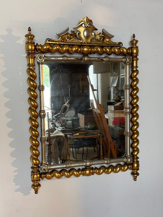 Specchiera antica dallo stile barocco, caratterizzata da una sontuosa cornice in legno intagliato con finitura dorata e dettagli argentati.