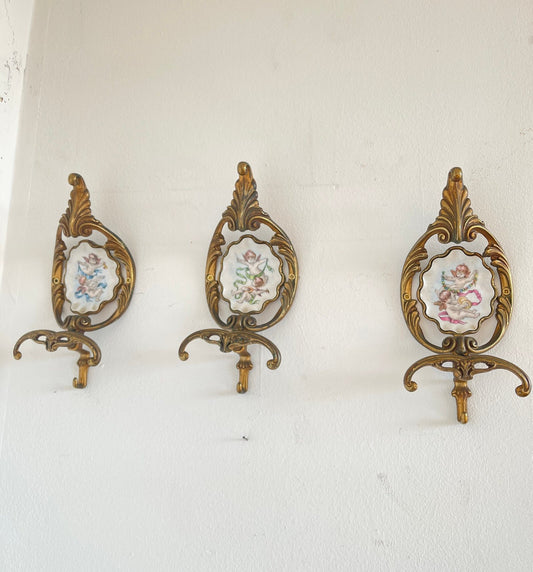 Set di 3 appendiabiti vintage in ottone con porcellana dipinta a mano - stile barocca Raffinato set di tre appendiabiti da parete in stile barocca, realizzati in ottone lavorato con inserti in porcellana dipinta a mano. Ogni pezzo presenta una decorazion